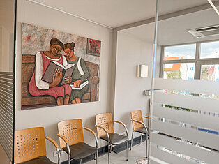 Wartebereich mit Gemälde im MVZ Goldbach Heller Wartebereich im MVZ Goldbach mit Holzstühlen und einem Gemälde zweier lesender Frauen an der Wand.