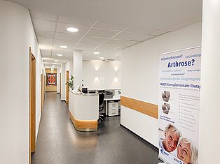 Empfangsbereich mit Infostand zur Arthrosetherapie im MVZ Goldbach Heller Flur mit Empfangstresen im MVZ Goldbach, Infoposter zur MBST-Therapie und moderner Wandgestaltung.