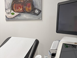 Detailansicht Ultraschallraum im MVZ Goldbach Nahaufnahme einer Behandlungsliege und eines Ultraschallgeräts im MVZ Goldbach mit Gemälde einer Arzttasche an der Wand.